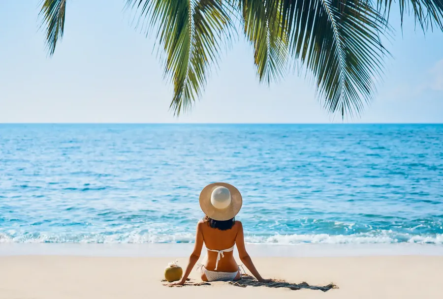 Palmiana de espaldas, con sombrero de paja en una playa tropical bajo una hoja de palmera